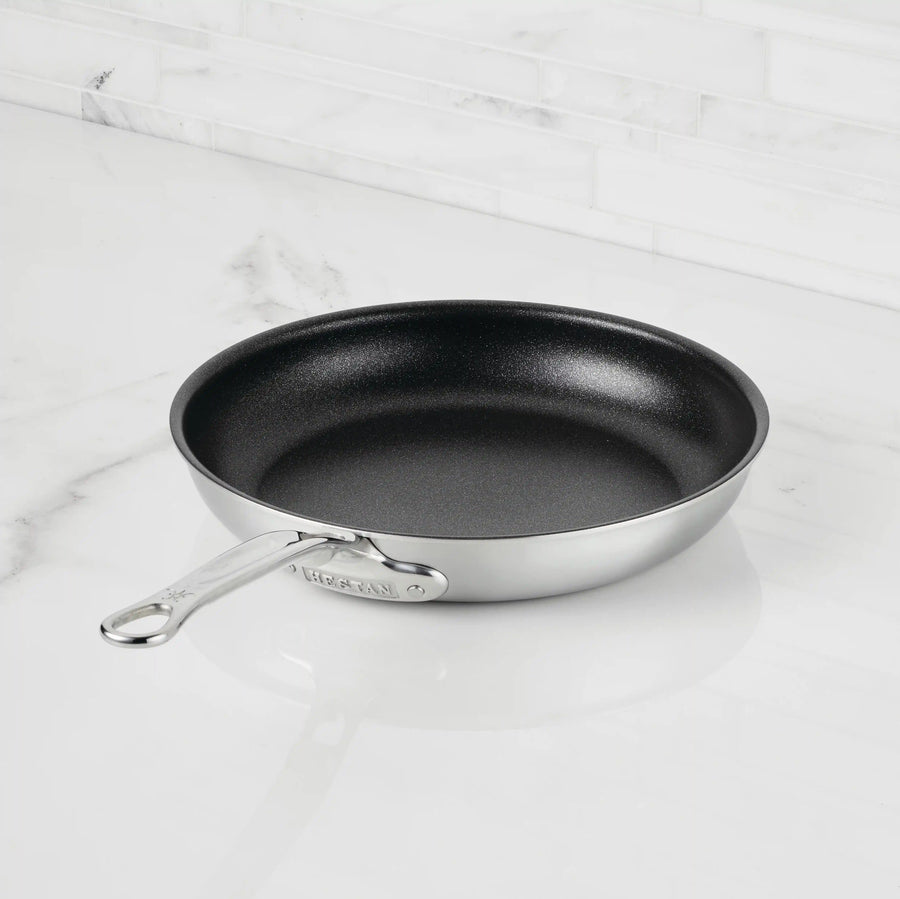 A Thomas Keller Insignia Commercial Clad Stainless Steel TITUM® Nonstick Skillet with a metal handle rests on a white marble countertop, backed by a white tile backsplash.