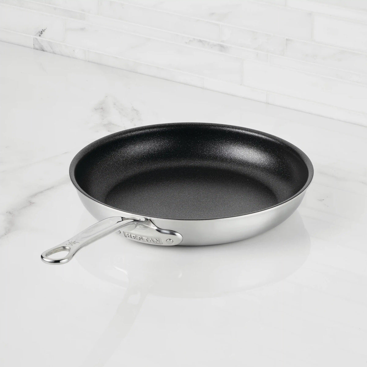 The Thomas Keller Commercial Clad Stainless Steel TITUM® Nonstick Skillet sits on a white marble countertop before a tiled backsplash, delivering professional culinary performance to your kitchen.