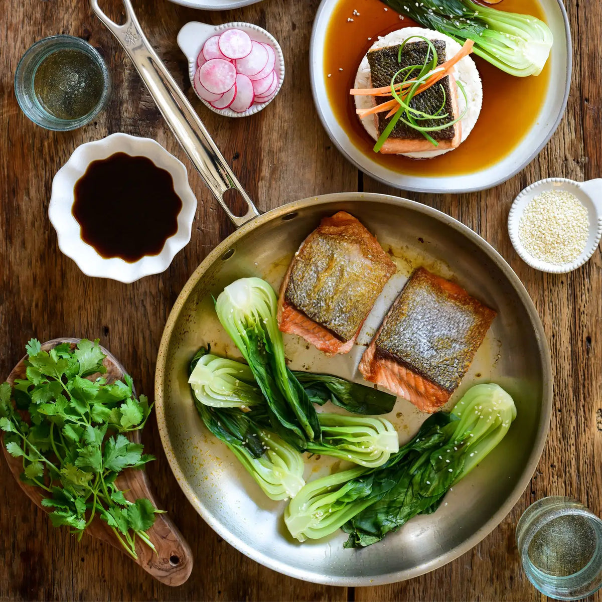 A wooden table set with a Thomas Keller Commercial Clad Stainless Steel Skillet of cooked salmon and bok choy, a fish plate with sauce, soy sauce bowl, sliced radishes, sesame seeds, cilantro, tongs, and two water glasses.