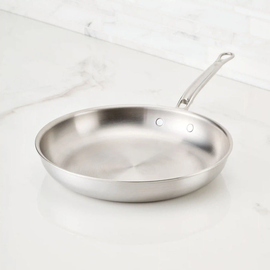 A Thomas Keller Insignia Commercial Clad Stainless Steel Skillet with an aluminum base and long handle sits empty on a white marble countertop, set against a backdrop of white tiled wall.