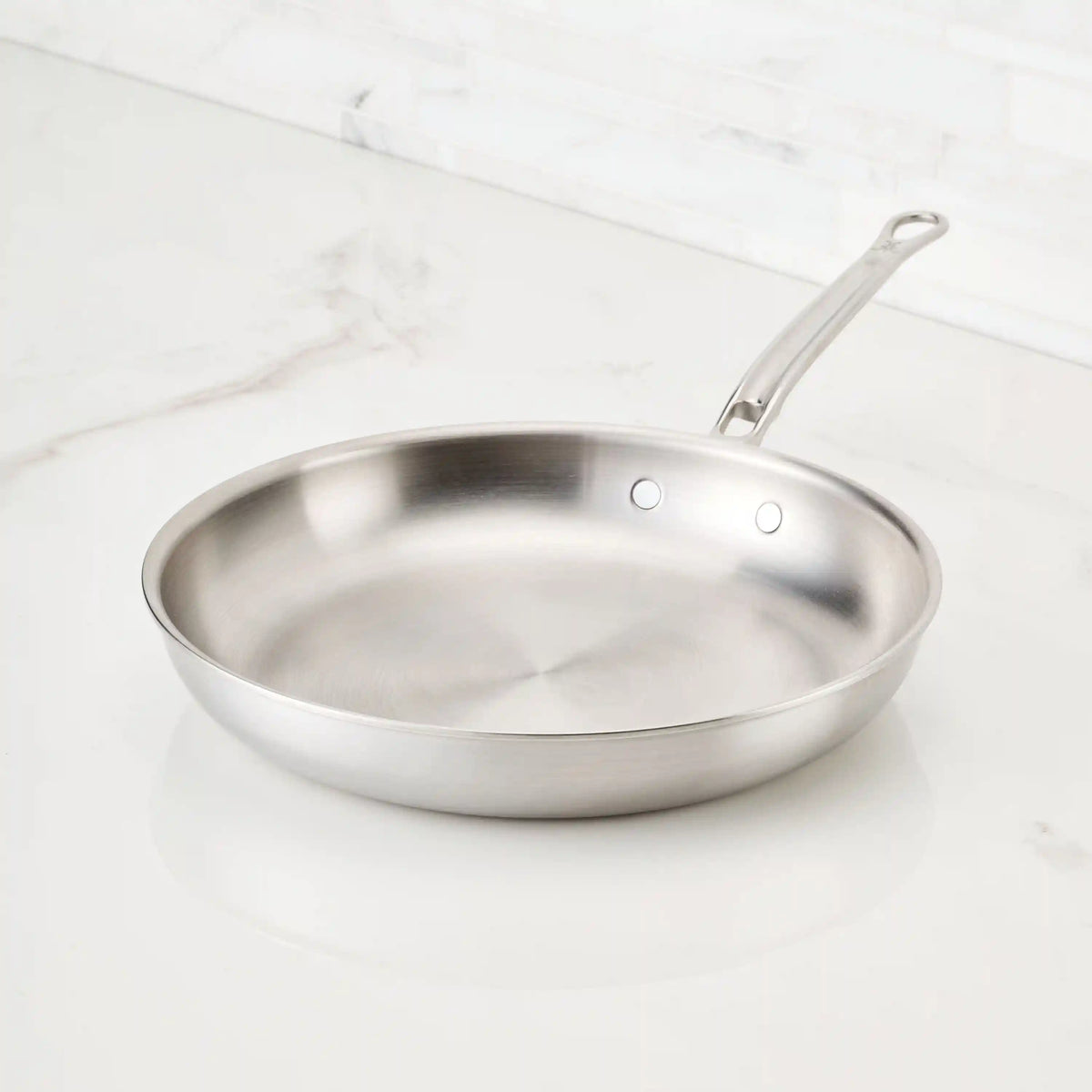 A Thomas Keller Commercial Clad Stainless Steel Skillet with a metal handle sits on a white marble countertop in a bright kitchen, showcasing the professional quality of Stainless Steel Skillets.