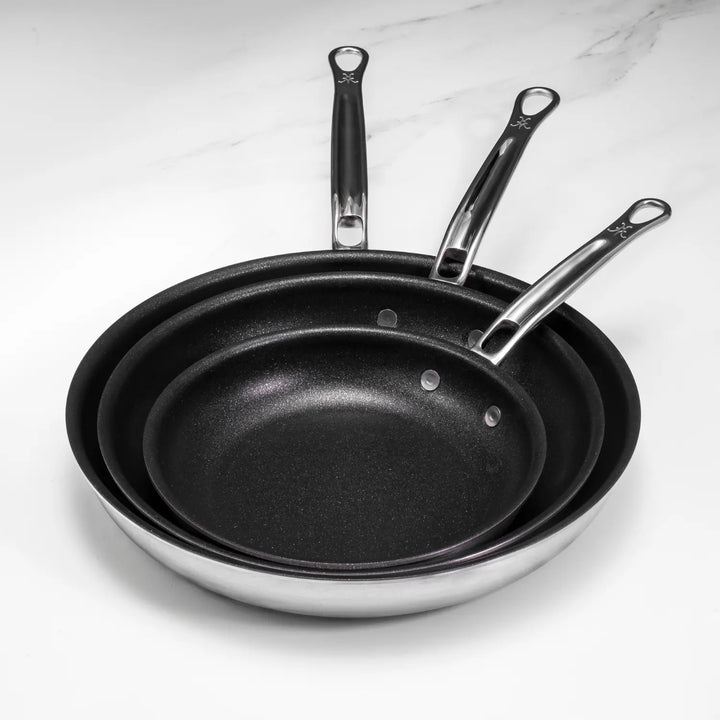 The Thomas Keller Insignia Commercial Clad Stainless Steel TITUM® Nonstick Skillets, nested in three sizes for professional use, feature silver handles with a small logo near the grips. They are elegantly displayed on a light marble surface.