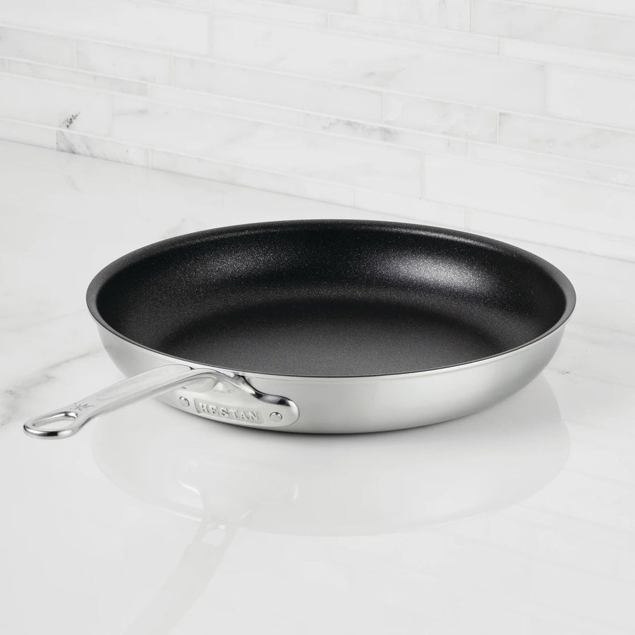 The Thomas Keller Insignia Commercial Clad Stainless Steel TITUM® Nonstick Skillet sits on a white marble countertop with a white tile backsplash.