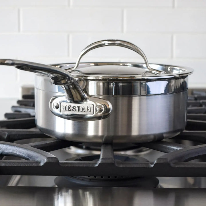 A ProBond stainless steel saucepan from the Professional Clad Ultimate 10-piece set sits on a black gas stove, its handle embossed with the brand name. The stove stands against white subway tiles.