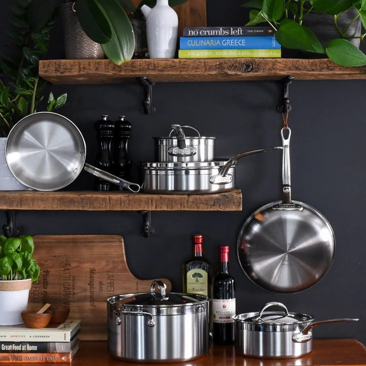 A kitchen scene features a ProBond Professional Clad Stainless Steel Covered Sauté Pan with a tri-ply body on wooden shelves. Culinaria Greece and no crumbs left cookbooks sit above olive oil and balsamic vinegar bottles, while green plants enhance the natural ambiance.