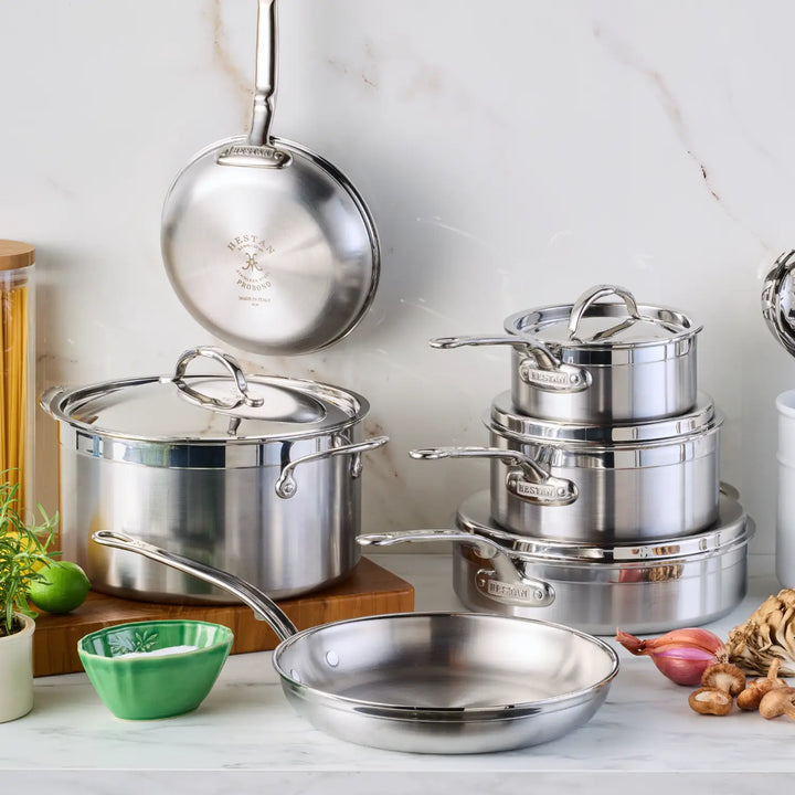 The ProBond Professional Clad 10-piece Stainless Steel Ultimate Set, featuring a frying pan, saucepans with lids, and a stockpot, is displayed on a marble countertop alongside a small green bowl, wooden containers, and vegetables.