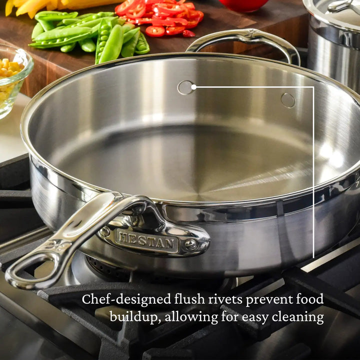 A stainless steel cooking pan from ProBond’s 10-piece Professional Clad set sits on a stovetop, exhibiting its chef-designed flush rivets and Tri-Ply Body for effortless cleaning and superior heat distribution. Sliced vegetables linger in the background.