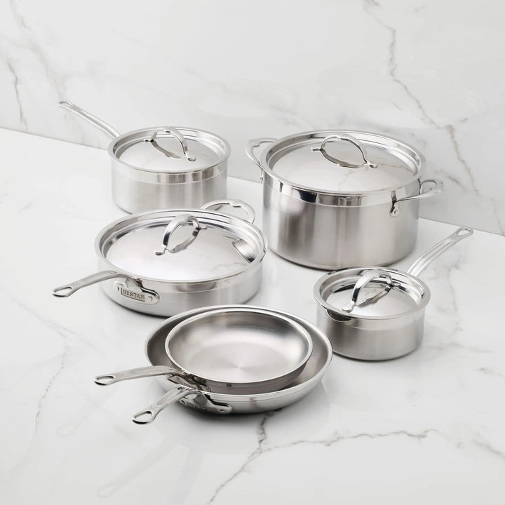 The ProBond® Forged™ Brushed Clad Stainless Steel Ultimate Set, 10-piece, handcrafted in Italy, features pots and pans with lids—saucepans, frying pans, and a stock pot—displayed on a white marble countertop with a marble backsplash.
