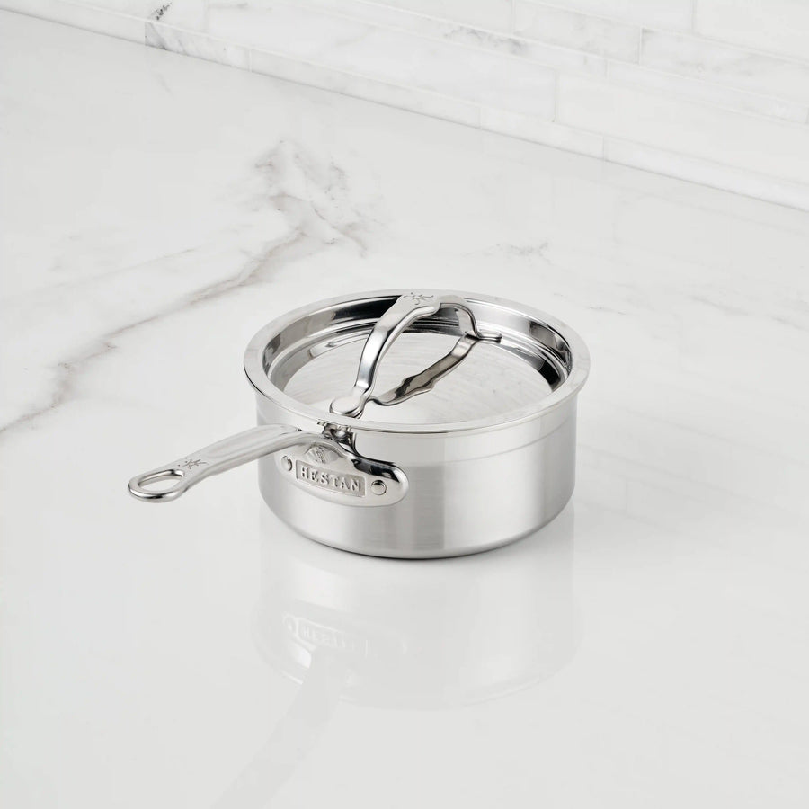 A ProBond® Forged™ Professional Clad Stainless Steel Saucepan with tri-ply body and aluminum core, featuring a long handle, polished finish, and lid, sits on a white marble countertop with gray veining.