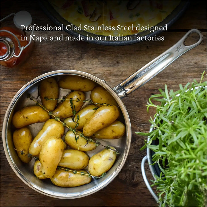 A ProBond Professional Clad Stainless Steel Saucepan features cooked yellow potatoes with herbs. A green leafy plant and sauce jar complete the scene. Made in Italy, this tri-ply aluminum core saucepan is designed in Napa.