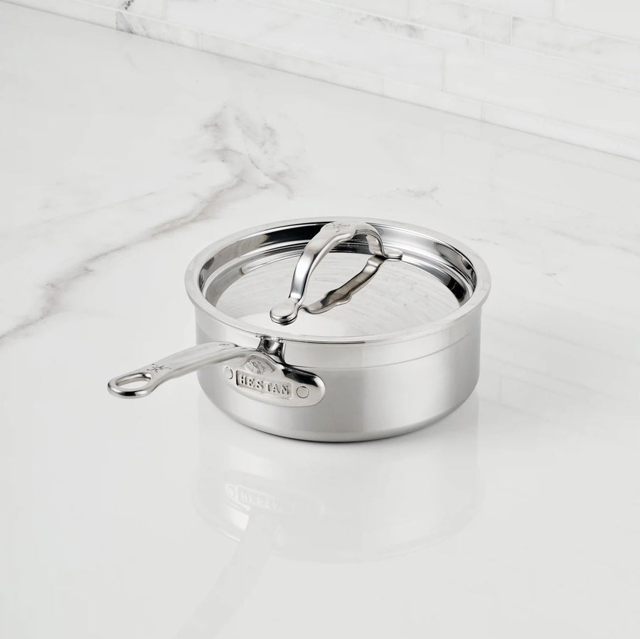 A ProBond® Forged™ Professional Clad Stainless Steel Saucepan with a tri-ply body and lid rests on a white marble countertop, featuring a long handle, sturdy modern design, and positioned before a white tiled backsplash.