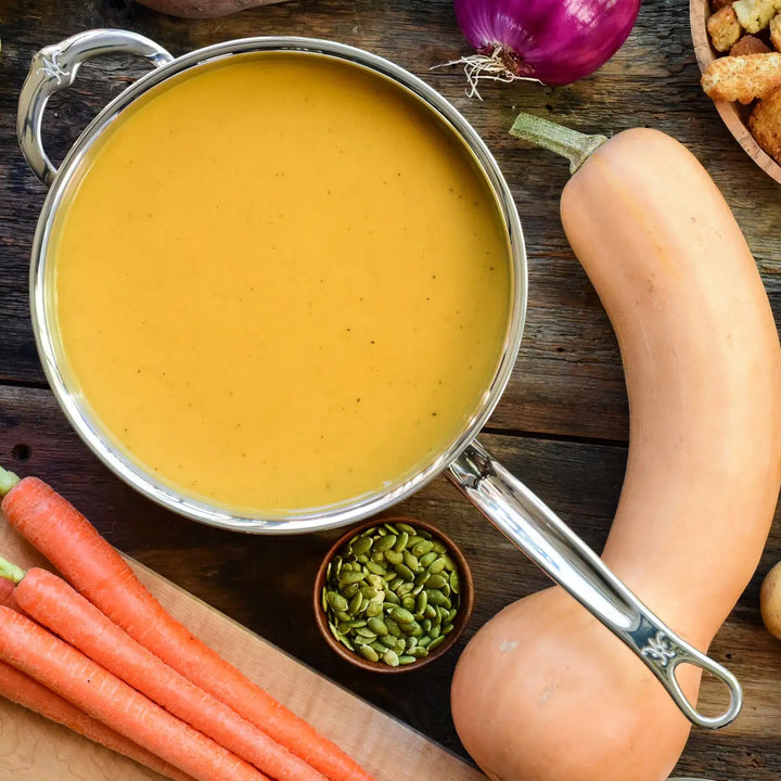 A pot of orange soup simmers in a ProBond Professional Clad Stainless Steel Saucepan on a wooden table, surrounded by raw butternut squash, carrots, and pumpkin seeds. Nearby are diced bread pieces and a red onion, ready to complement the culinary masterpiece crafted with this cookware.