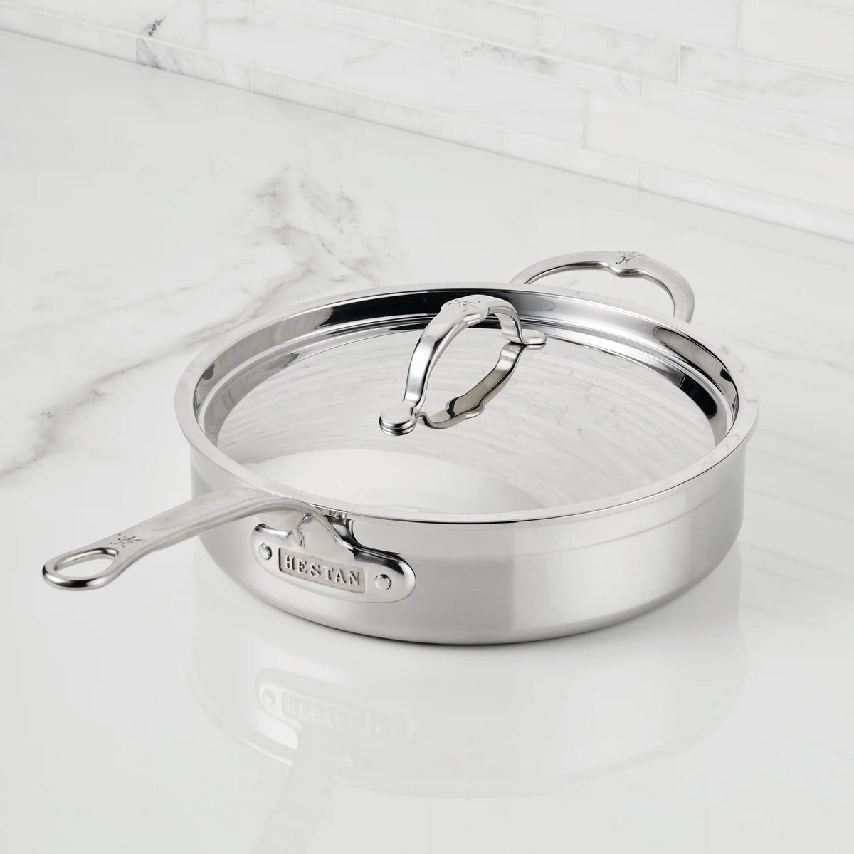 A ProBond® Forged™ Professional Clad Stainless Steel Covered Sauté Pan with two handles and a lid rests on a white marble countertop, backed by a marble backsplash—ideal for any professional kitchen.