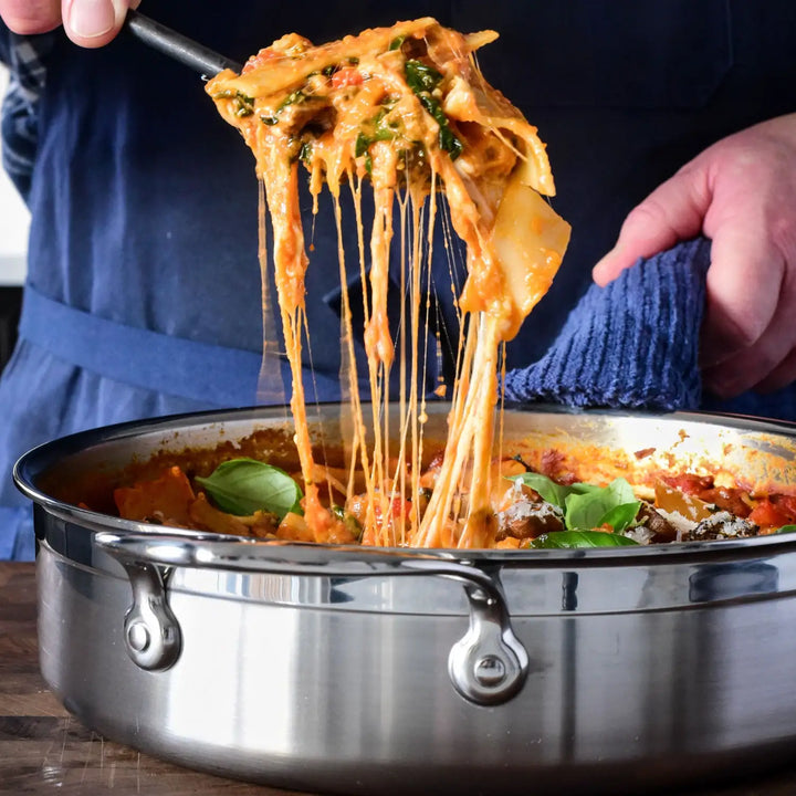 Wearing a blue apron, a person ladles cheesy lasagna from a ProBond Professional Clad Stainless Steel Covered Sauté Pan. Overflowing with melted cheese, pasta, vegetables, and fresh basil, the dish contrasts with the dark blue cloth highlighting the pans sleek tri-ply body.