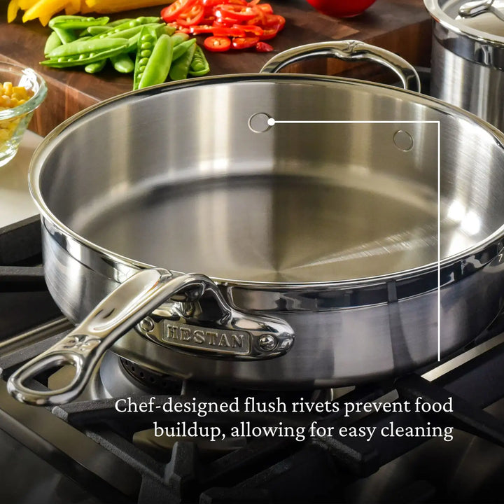 A ProBond Professional Clad Stainless Steel Covered Sauté Pan with flush rivets sits on a stovetop, featuring red and green chopped peppers with a spoon in the background. The design prevents food buildup for effortless cleaning.