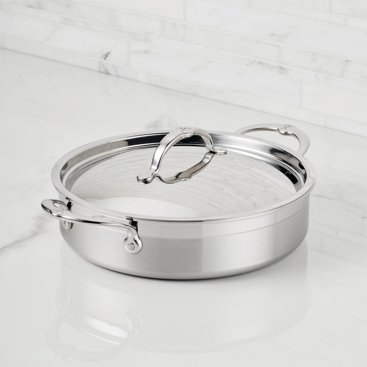 A ProBond® Forged™ Professional Clad Stainless Steel Braiser with Lid, 3.5-Quart, featuring two handles, sits on a white marble countertop against a white tiled backsplash.