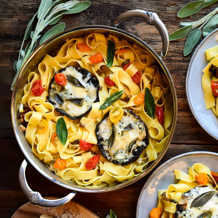 The ProBond Professional Clad Stainless Steel Braiser with Lid, 3.5-Quart, holds tagliatelle pasta with cherry tomatoes and sage leaves, topped with large stuffed mushroom slices. It rests on a rustic wooden surface surrounded by green sprigs, showcasing culinary elegance.