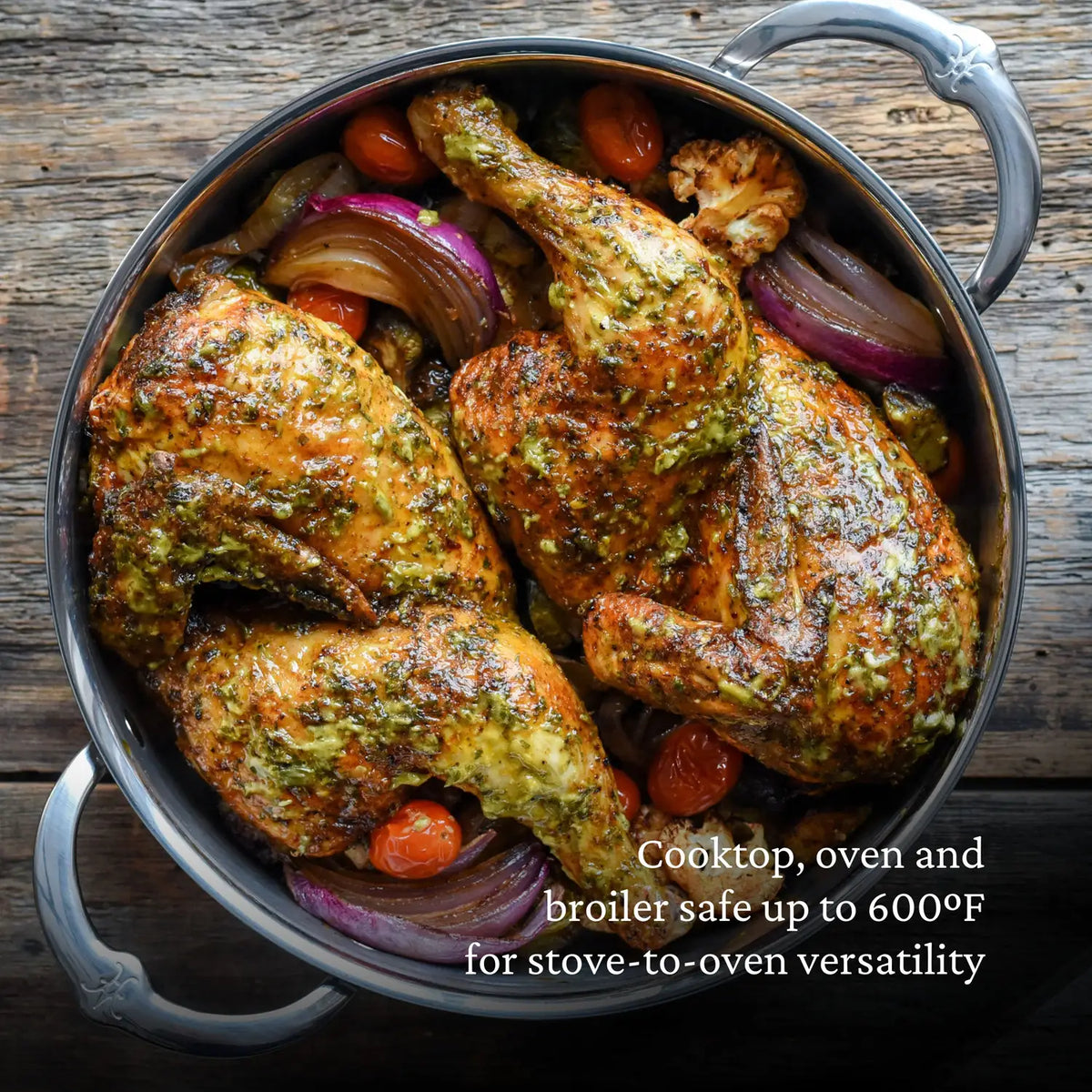 A juicy cooked chicken sits in a 3.5-quart ProBond Professional Clad Stainless Steel Sauteuse, seasoned with herbs and spices. Roasted red onion wedges and cherry tomatoes surround it on the wooden surface. A text overlay reads, Cooktop, oven, and broiler safe up to 600°F. Handcrafted in Italy.