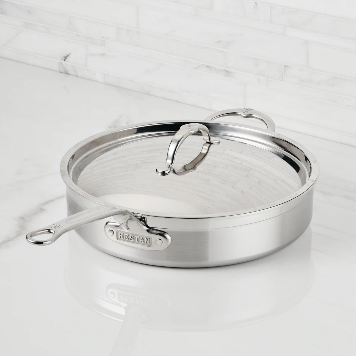 A ProBond® Forged™ Professional Clad Stainless Steel Covered Sauté Pan, with a lid and long handle, rests on a white marble countertop with a matching marble backsplash.