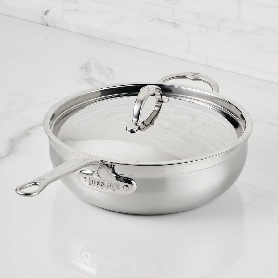 A ProBond® Forged™ Professional Clad Stainless Steel Essential Pan with Tri-Ply Construction, long and helper handles, sits on a white marble countertop with its matching lid propped against the rim.