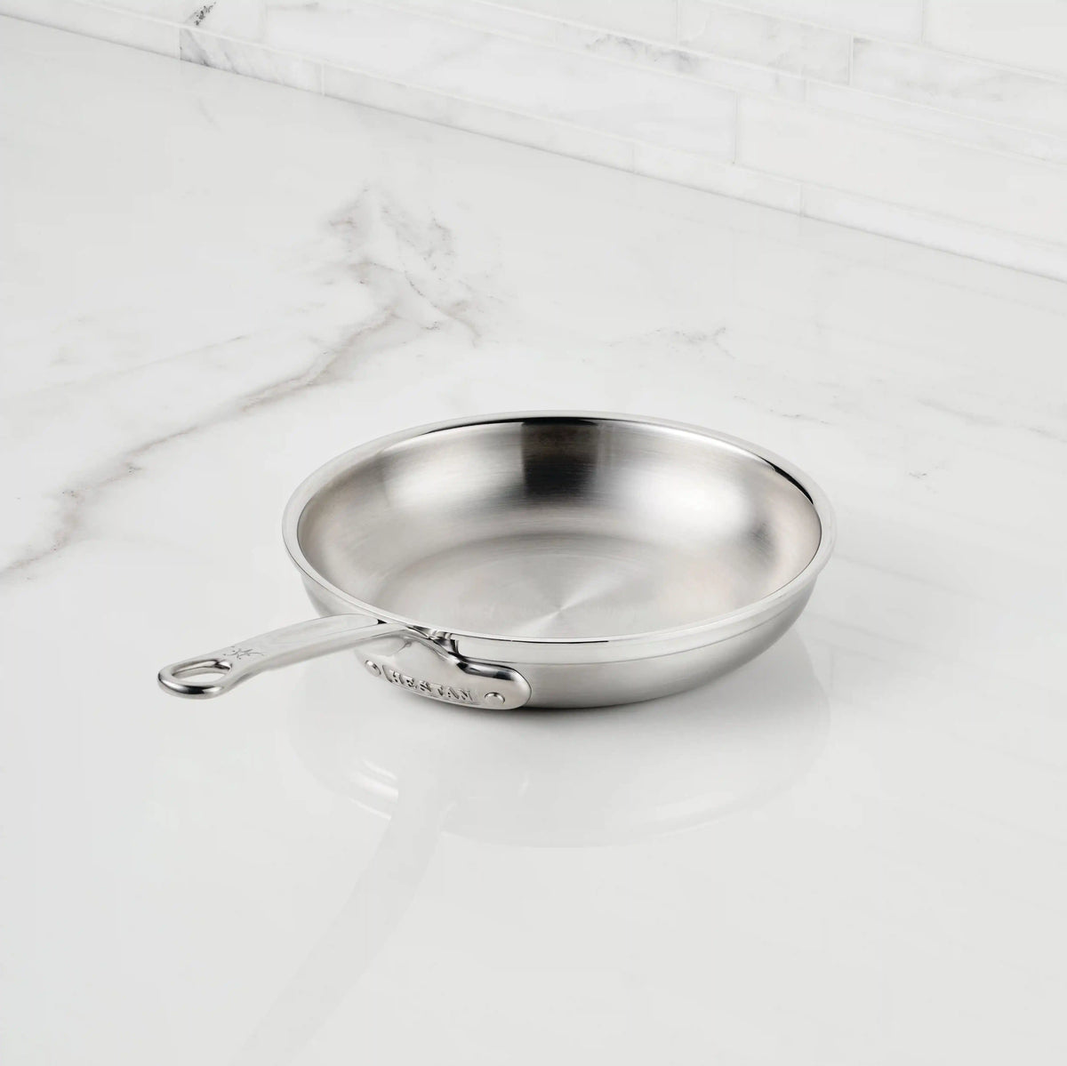 The ProBond® Forged™ Professional Clad Stainless Steel Skillet, with a tri-ply body and metal handle, sits on a white marble countertop. Its polished surface reflects light, ensuring fast, even heat distribution for efficient cooking.
