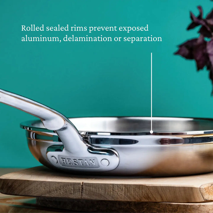 A ProBond Professional Clad Stainless Steel Skillet with a tri-ply body and ergonomic handle sits on a wooden board against a teal background. Text above notes the rolled sealed rims that prevent exposed aluminum, delamination, or separation. A plant is partially visible on the right.