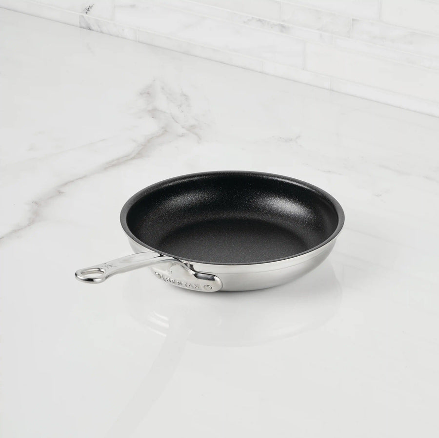 A ProBond® Forged™ Professional Clad Stainless Steel TITUM® Nonstick Skillet with a black interior and metal handle sits on a white marble countertop against a tiled backsplash.
