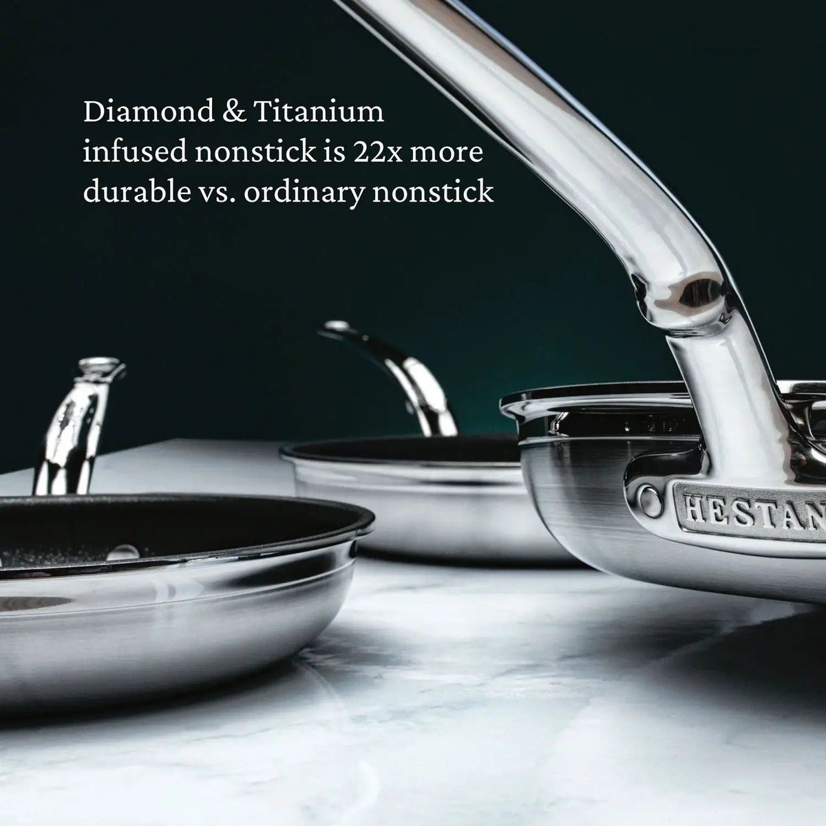 Close-up of three stainless steel pans, including a ProBond Professional Clad Stainless Steel TITUM® Nonstick Skillet, with shiny handles on marble. Text atop reads, Diamond & Titanium infused nonstick is 22x more durable vs. ordinary nonstick. The brand name HESTAN appears on the handle.