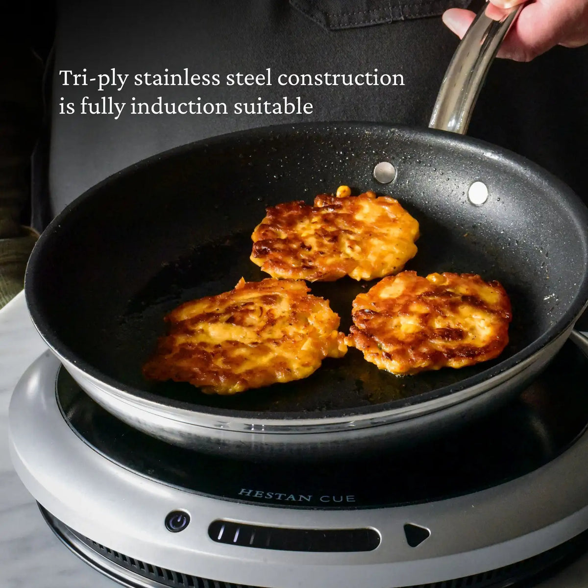A person holds a ProBond Professional Clad Stainless Steel TITUM® Nonstick Skillet with three golden-brown fritters inside, placed on an induction cooktop. The text reads, Clad stainless steel construction is perfect for induction cooking.