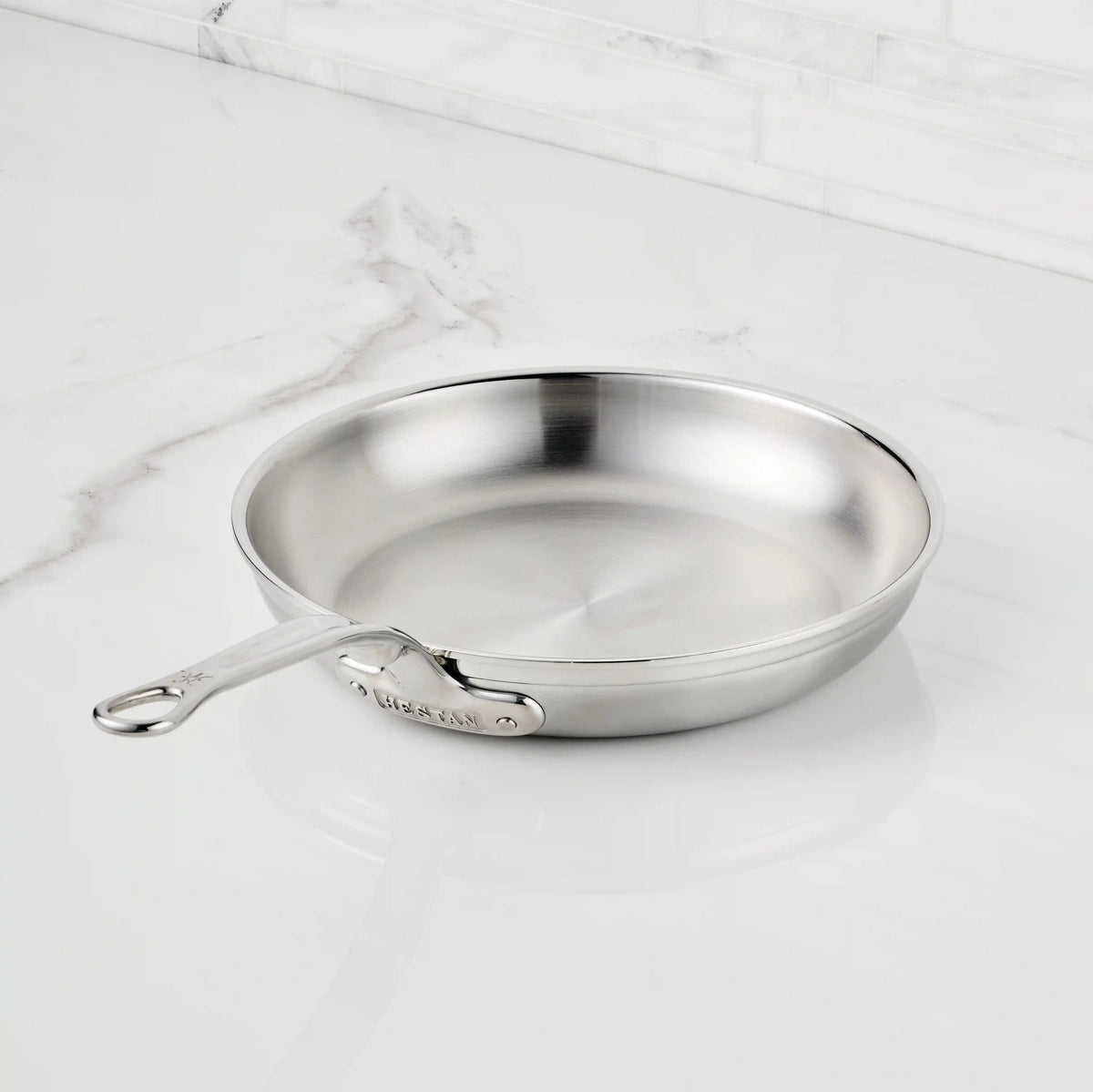 A ProBond® Forged™ Professional Clad Stainless Steel Skillet with a tri-ply body and long handle rests on a white marble countertop, complemented by a matching marble backsplash.