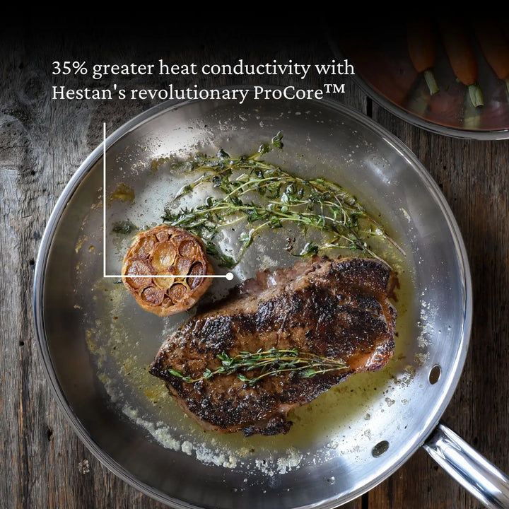 A steak sizzles in the ProBond Professional Clad Stainless Steel Skillet, surrounded by herbs and a halved garlic bulb. The top text highlights 35% greater heat conductivity with Hestans ProCore™ technology, boasting ergonomic handles and a tri-ply body for perfect results.