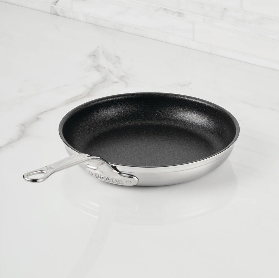 A ProBond® Forged™ Professional Clad Stainless Steel TITUM® Nonstick Skillet, handcrafted in Italy and featuring a long handle with slightly rounded edges, rests on a white marble countertop.