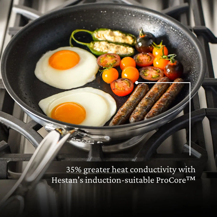 A frying pan on a stovetop showcases two sunny-side-up eggs, grilled vegetables, and sausages. Text overlay reads, 35% greater heat conductivity with ProBonds induction-suitable Professional Clad Stainless Steel TITUM® Nonstick Skillets.