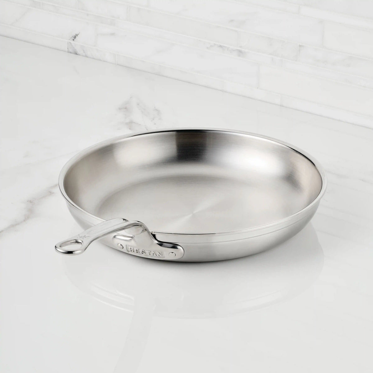 A ProBond® Forged™ Professional Clad Stainless Steel Skillet with tri-ply construction and a sturdy handle rests on a white marble countertop with a matching backsplash in the background.