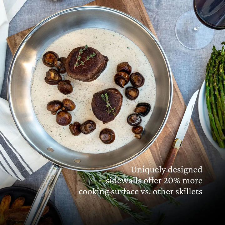 A ProBond Professional Clad Stainless Steel Skillet with a tri-ply body holds two pieces of meat topped with herbs and mushrooms in cream sauce, resting on a wooden board next to cutlery, red wine, and asparagus. The skillet is known for its ergonomic handles. Text overlays the image.