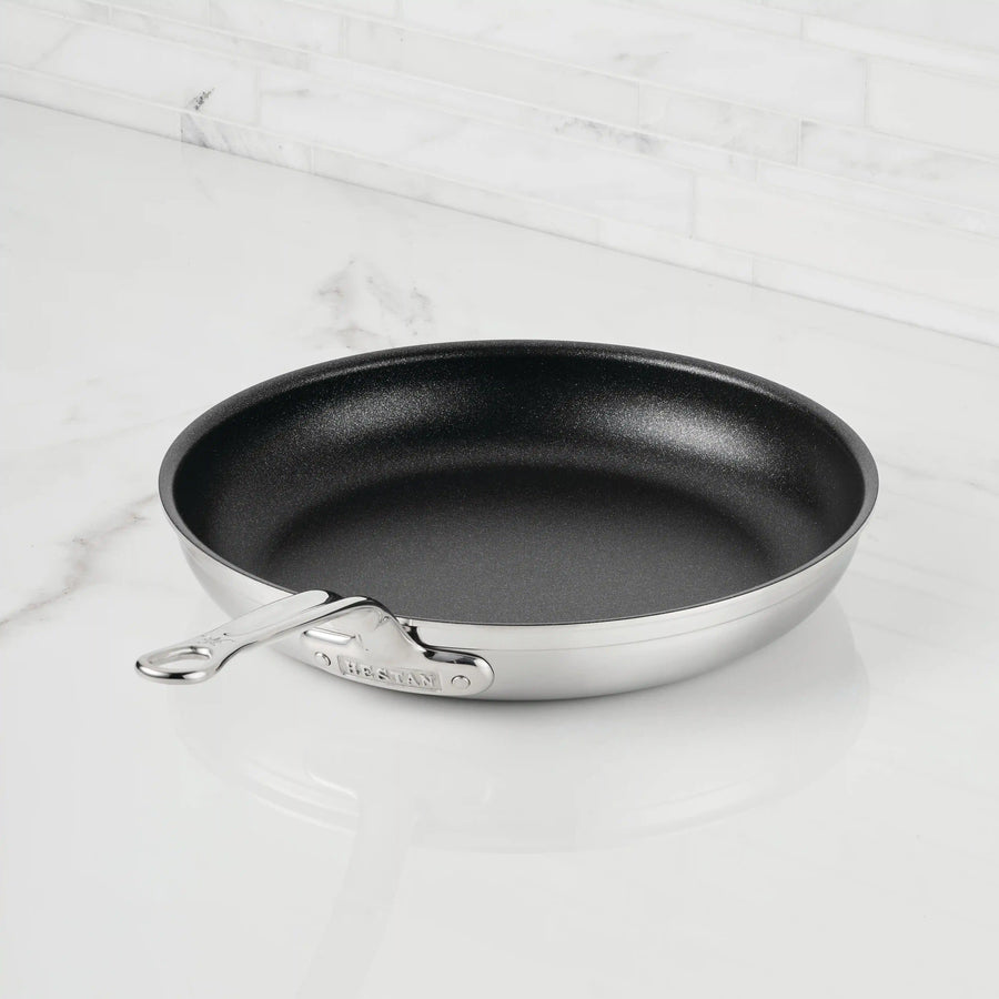 A ProBond® Forged™ Professional Clad Stainless Steel TITUM® Nonstick Skillet with a sleek metal handle sits on a white marble countertop, backed by a crisp white tile backsplash.