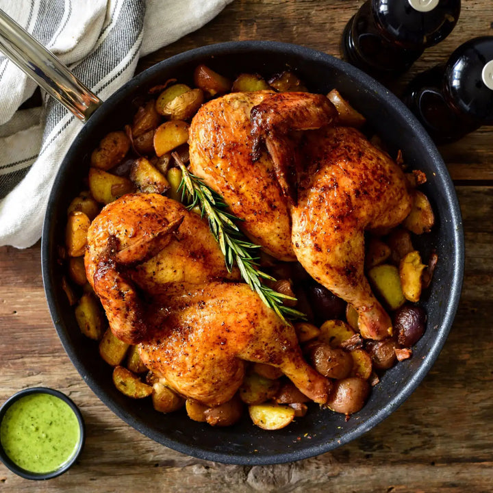 A top view of two roasted chicken halves in a ProBond Professional Clad Stainless Steel TITUM® Nonstick Skillet, surrounded by potatoes, with rosemary on top. A striped cloth is nearby alongside a small bowl of green sauce and two pepper grinders on a wooden surface.