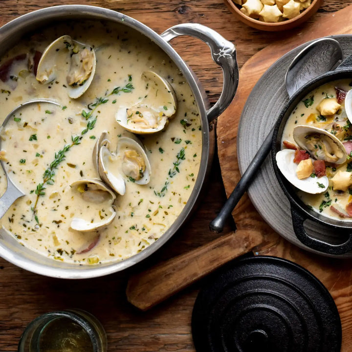 A pot of creamy clam chowder sits on a wooden surface in a ProBond Professional Clad Stainless Steel Soup Pot, 3-Quart. Its tri-ply body is revealed as a ladle rests inside, next to a black bowl of steaming chowder with crackers, and a small plate with more crackers alongside.