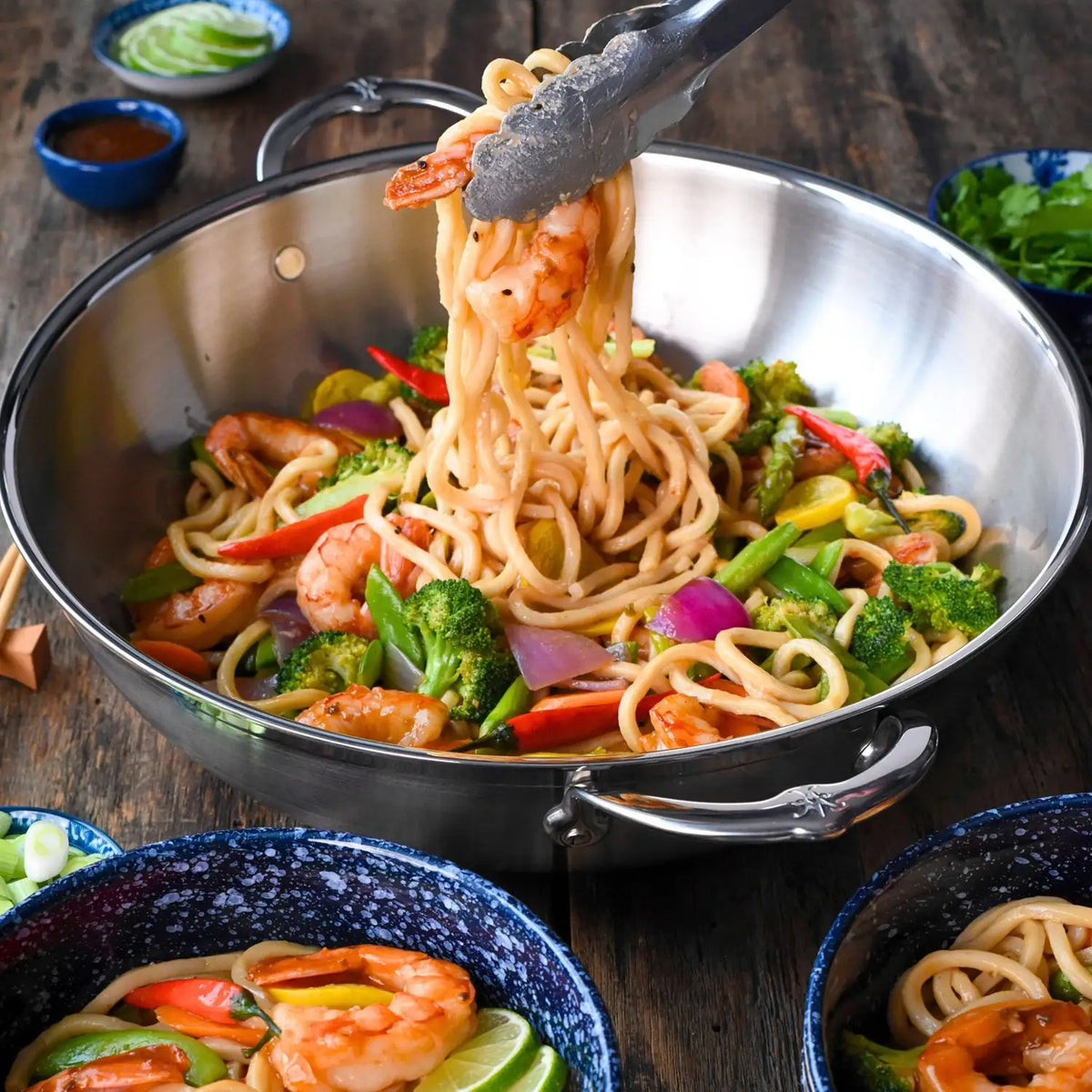 A bowl of shrimp stir-fried noodles with broccoli, red peppers, and onions is served using tongs from a ProBond Professional Clad Stainless Steel Wok, 14-Inch on a wooden table. Nearby are small bowls with sliced cucumbers, lime wedges, and sauce for added flavor.