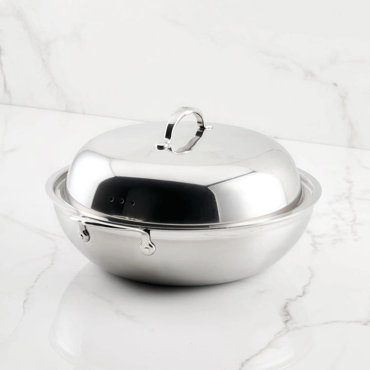 The ProBond® Forged™ Brushed Clad Stainless Steel Wok with Dome Lid, 14-Inch, featuring two side handles and a reflective tri-ply design for even heat, is shown on a white marble surface.