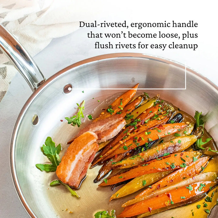 The CopperBond Copper Induction Skillet elegantly holds salmon fillets, roasted carrots, and greens on a white surface. With a stainless steel design and copper core, its dual-riveted ergonomic handle ensures durability and easy cleanup. Induction compatible.