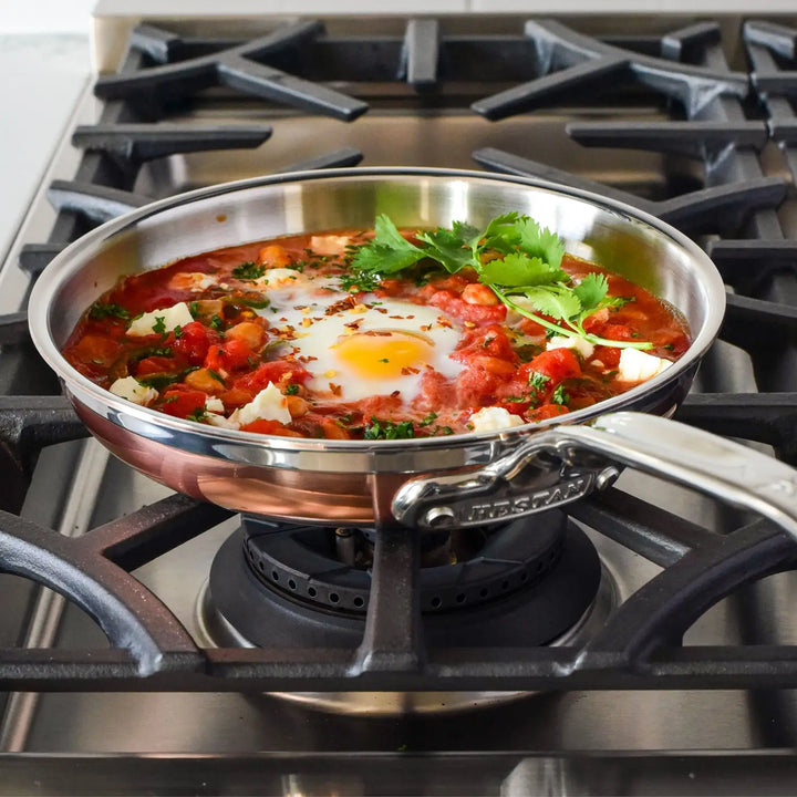 A CopperBond Copper Induction Skillet with 5-layer construction cradles a vibrant dish of tomatoes, poached eggs, and herbs, possibly cilantro. The eggs nestle in a red sauce containing cheese or vegetable chunks, ensuring even cooking on any stovetop.