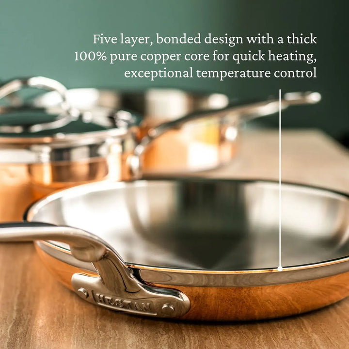 The CopperBond Copper Induction Skillet, with its stainless steel exterior and induction-compatible design, elegantly rests on a wooden surface. A five-layer bonded system with a copper core ensures quick heating, while the pot with lid in the background emphasizes its allure.