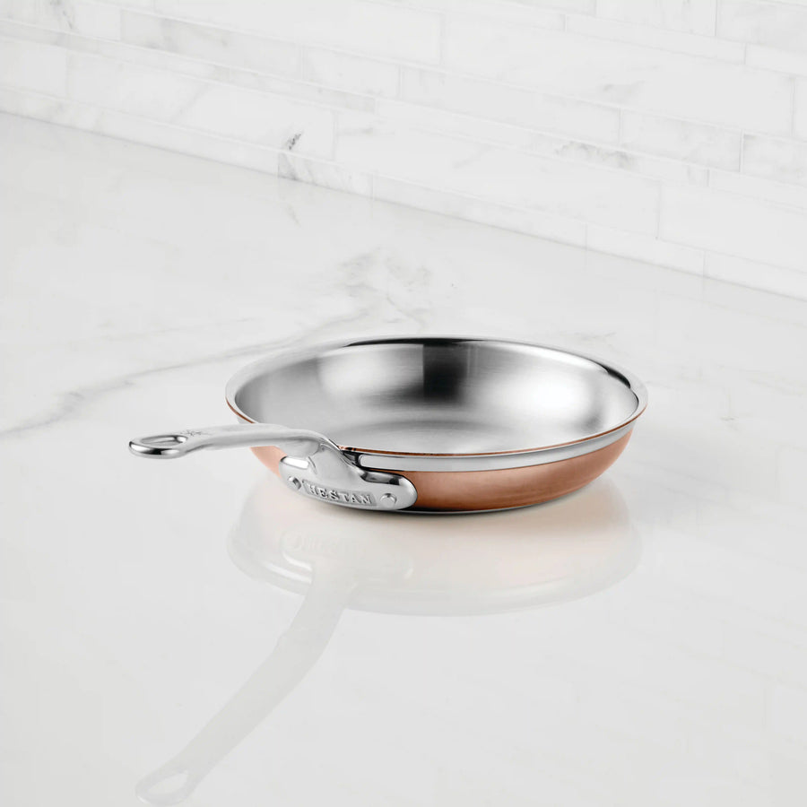 A CopperBond Copper Induction Skillet with a stainless steel body and metal handle sits on a white marble countertop in a kitchen with a white tiled backsplash, highlighting the sophistication of CopperBond cookware.