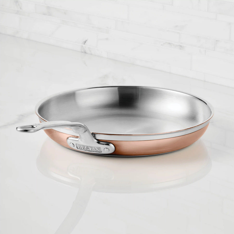 A CopperBond Copper Induction Skillet with a 5-layer stainless steel interior and metal handle sits on a white countertop in front of a white tiled backsplash.