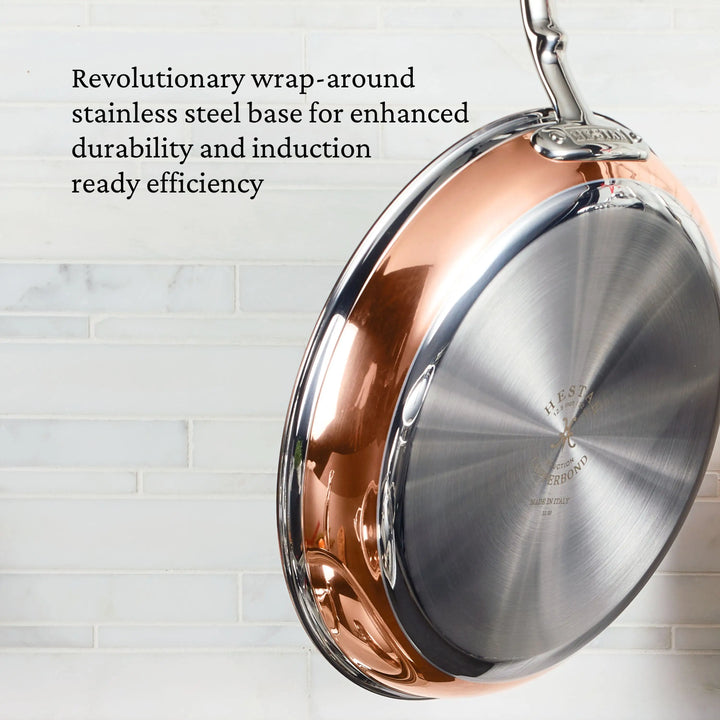 A close-up of a CopperBond Copper Induction Ultimate Set 10-Piece frying pan hanging on a wall, with text noting its stainless steel base for durability and induction efficiency. White tile backsplash in the background.