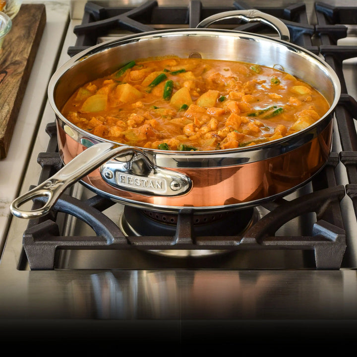 A CopperBond Copper Induction Ultimate Set pan with a stainless steel handle rests on a gas stove, holding thick orange stew filled with meat, potatoes, and green vegetables.