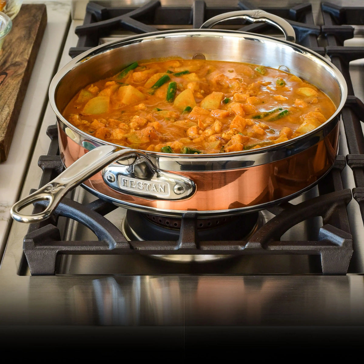 A CopperBond Copper Induction Ultimate Set pan with a stainless steel handle rests on a gas stove, holding thick orange stew filled with meat, potatoes, and green vegetables.