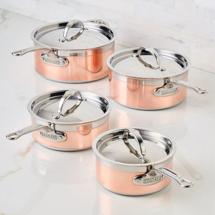 Four CopperBond copper and stainless steel induction saucepans sit on a white marble surface. Each pot has a metal handle and bears the brand name. A light-colored tile wall serves as the background.