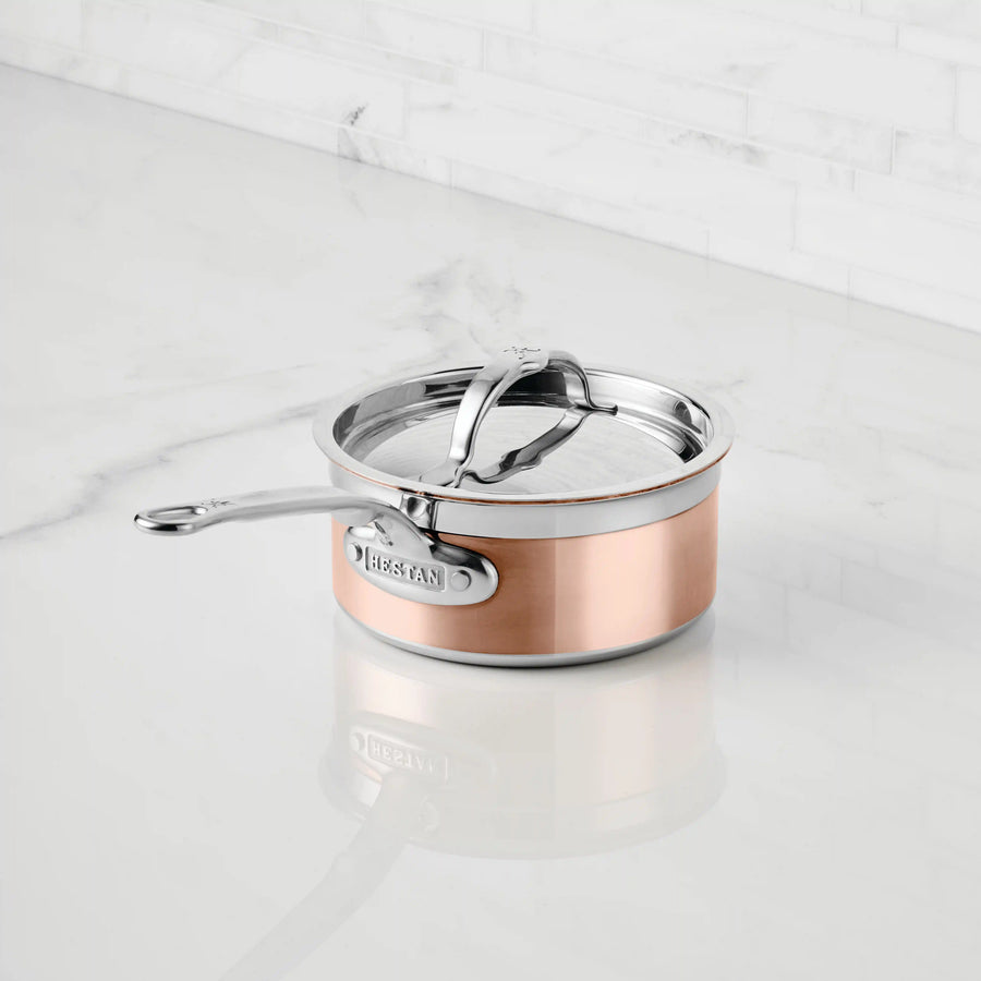 A CopperBond Copper Induction Saucepan with a stainless steel handle and lid sits on a white marble countertop, with a marble backsplash in the background.